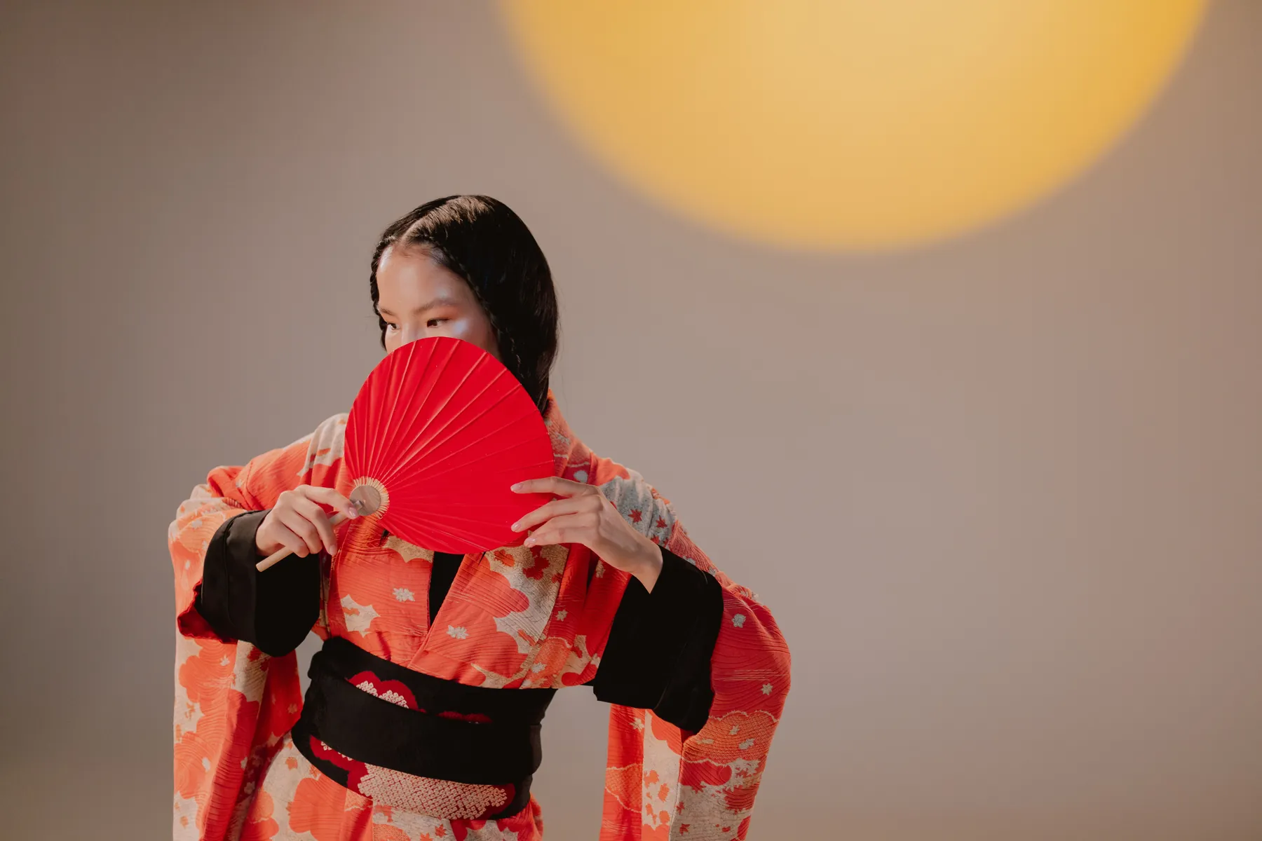 En kvinde i en orange og sort kimono holder en rød foldevifte foran ansigtet i varmt studiolys