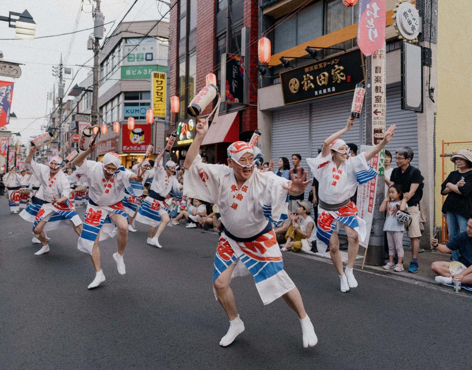 Dansere i hvide og blå happi-frakker bevæger sig ned ad en afspærret gade ved en japansk festival, mens tilskuere ser til fra kantstenen